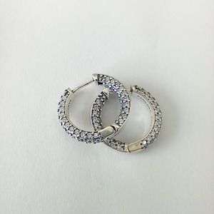 May include: A pair of silver hoop earrings encrusted with small, clear gemstones. The earrings are round and feature a hinged closure. The brand name "Pandora" is visible on the side of one earring.