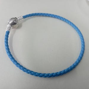 Könnte beinhalten: Ein hellblaues geflochtenes Lederarmband mit einem silberfarbenen Verschluss. Das Armband ist kreisförmig und scheint ungetragen zu sein. Der Verschluss hat ein dekoratives Design. Es ist ein Schmuckstück.