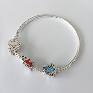 Könnte beinhalten: Silbernes Charm-Armband mit drei farbenfrohen Charms. Die Charms umfassen ein Herz mit Text, eine rote Figur und eine blaue geflügelte Figur. Das Armband liegt auf einer weißen Oberfläche.