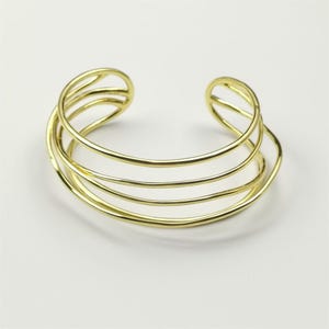 Peut inclure: Un bracelet manchette doré avec un design superposé. Le bracelet présente plusieurs bandes parallèles qui s'incurvent autour du poignet, créant un look ouvert et moderne. Le métal a une finition polie, reflétant la lumière.