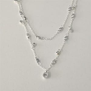 Puede incluir: Un delicado collar de plata en capas con un pequeño colgante. El collar presenta dos finas cadenas, adornadas con pequeñas cuentas redondas y un colgante en forma de lágrima con un diseño de corazón. La joyería se exhibe sobre un fondo blanco.