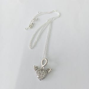 May include: A silver necklace with a heart-shaped pendant. The pendant is encrusted with small, clear stones and has small wings on either side. The chain is delicate and silver, with a clasp at the end.