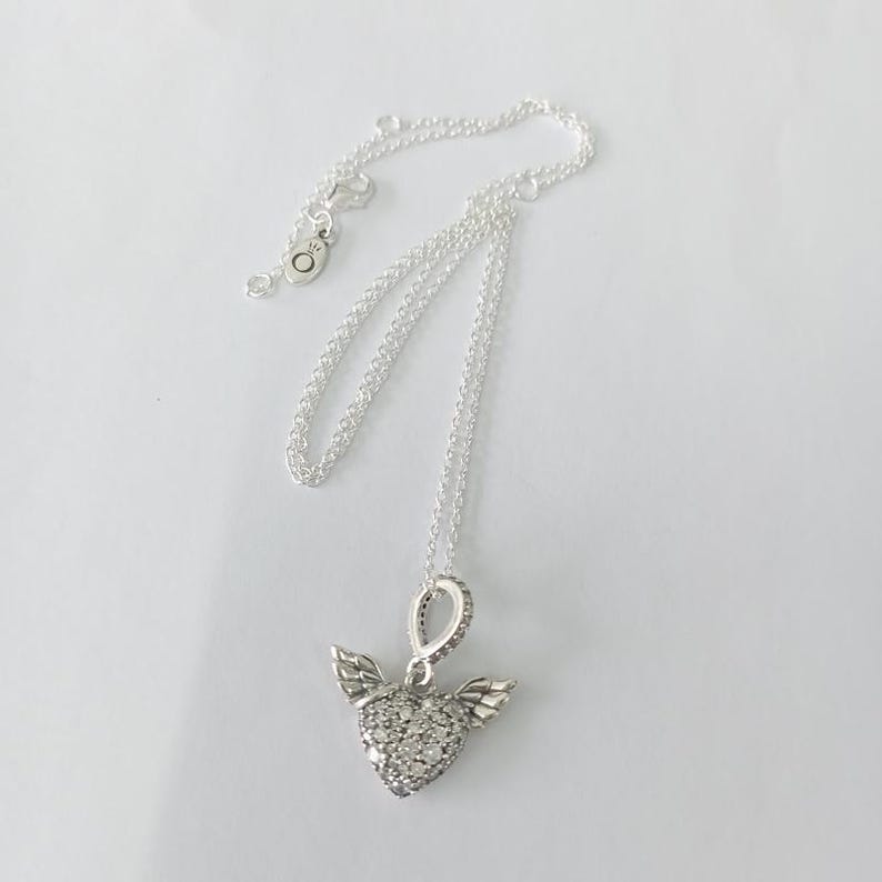 May include: A silver necklace with a heart-shaped pendant. The pendant is encrusted with small, clear stones and has small wings on either side. The chain is delicate and silver, with a clasp at the end.