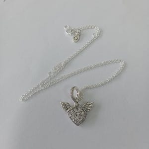 May include: A silver necklace with a heart-shaped pendant. The pendant is encrusted with small, clear stones and features small wings on either side. The necklace chain is delicate and silver.