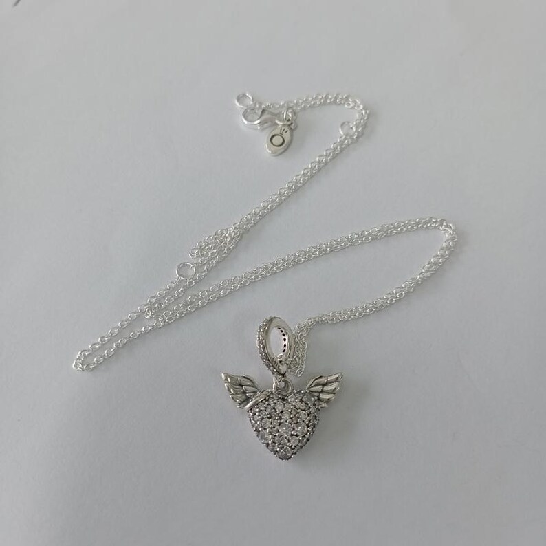 May include: A silver necklace with a heart-shaped pendant. The pendant is encrusted with small, clear stones and features small wings on either side. The necklace chain is delicate and silver.