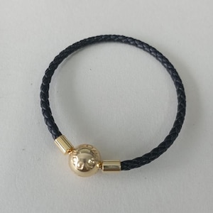 Puede incluir: Una pulsera de cuero trenzado negro con un cierre dorado. El cierre es una cuenta redonda con un diseño decorativo. La pulsera es circular y parece sin usar.