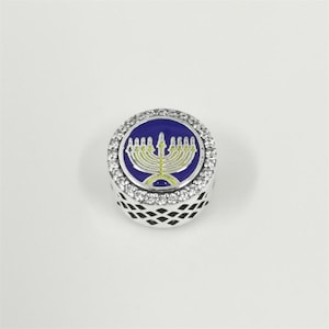 Op de afbeelding: Een zilverkleurige bedel met een blauw en geel menora-ontwerp. De ronde bedel heeft een ring van heldere kristallen rond het blauwe midden. De zijkanten van de bedel hebben een geometrisch patroon.