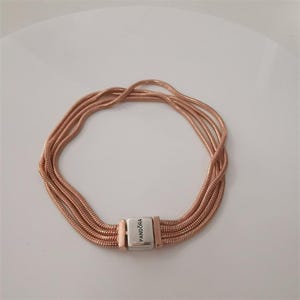 Könnte beinhalten: Roségoldenes Armband mit mehreren Strängen. Das Armband hat einen silbernen Verschluss mit dem eingravierten Markennamen "Pandora". Das Armband besteht aus einem glänzenden, metallischen Material.