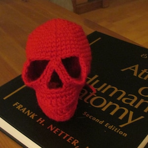 May include: A handmade, bright red crochet skull sits atop a book titled "Atlas of Human Anatomy." The skull's eye sockets and nasal cavity are clearly defined. The book's cover is black with gold lettering.