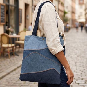 May include: A blue denim tote bag with a layered design. The bag has dark blue straps and is carried over the shoulder. The bag is filled with a book and a water bottle. The bag is being carried on a cobbled street.