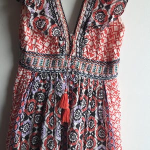 May include: A patterned summer dress with a V-neck and frill sleeves. The dress has a floral design in red, black, and purple, with a tassel at the waist. It is hanging on a white hanger. The dress is approximately 100 cm in length.