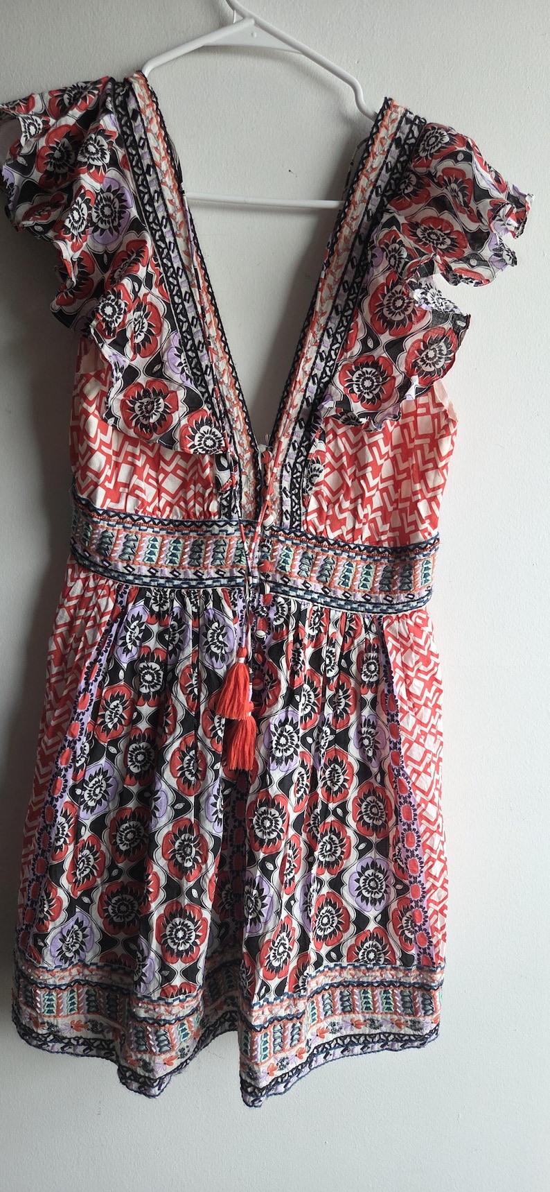 May include: A patterned summer dress with a V-neck and frill sleeves. The dress has a floral design in red, black, and purple, with a tassel at the waist. It is hanging on a white hanger. The dress is approximately 100 cm in length.