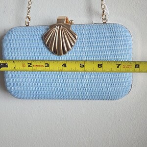 Puede incluir: Un bolso de mano rectangular de color azul claro con una superficie tejida texturizada y un cierre de metal dorado en forma de concha. Una correa de cadena dorada est&aacute; unida a la parte superior. Una cinta m&eacute;trica Stanley mide 20 cm de ancho.
