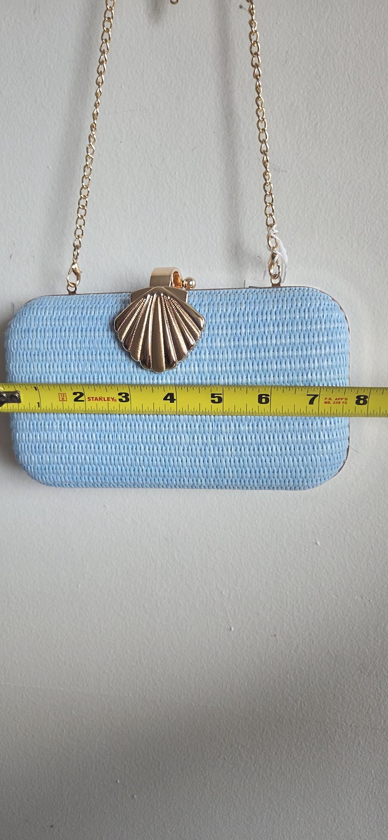 Puede incluir: Un bolso de mano rectangular de color azul claro con una superficie tejida texturizada y un cierre de metal dorado en forma de concha. Una correa de cadena dorada est&aacute; unida a la parte superior. Una cinta m&eacute;trica Stanley mide 20 cm de ancho.