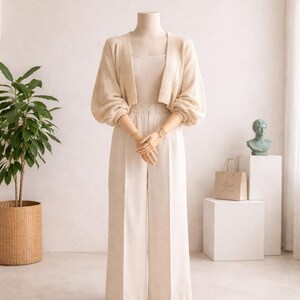 May include: A cream-colored outfit displayed on a mannequin. The ensemble includes wide-leg pants, a tank top, and a cropped cardigan with voluminous sleeves. A small paper shopping bag and a green bust statue are visible in the background.