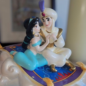 May include: A ceramic figurine of Aladdin and Jasmine riding on a magic carpet. The carpet is blue with gold trim and has a purple border. Aladdin is wearing a white turban and a gold sash. Jasmine is wearing a blue dress with a gold belt.
