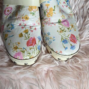 May include: Pair of white clogs with a floral pattern featuring pink, yellow, and blue flowers. The clogs have a wooden sole and a cream-colored strap. The brand name "Free People" is visible on the insole.