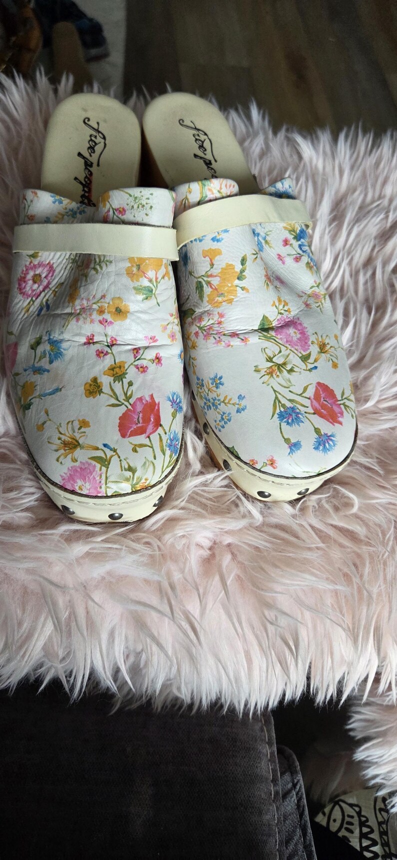 May include: Pair of white clogs with a floral pattern featuring pink, yellow, and blue flowers. The clogs have a wooden sole and a cream-colored strap. The brand name "Free People" is visible on the insole.