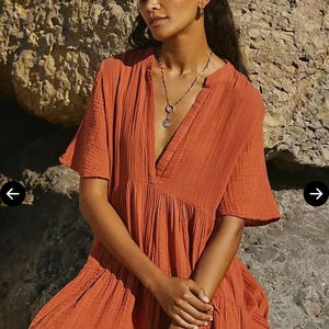 May include: Rust-colored mini dress with short sleeves and a V-neckline. The dress has a tiered skirt and is made of a textured fabric. The model is wearing a necklace and bracelet. The website editorialist.com is visible in the image.