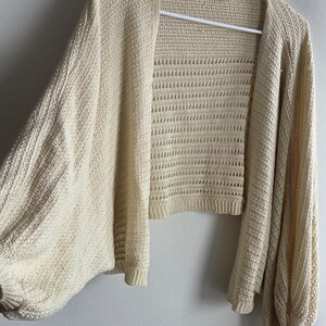 May include: Cream-colored open-front cardigan with long, voluminous sleeves. The knitwear features a textured pattern on the sleeves and a more open, airy weave on the body. The garment is hanging on a white hanger against a plain wall.