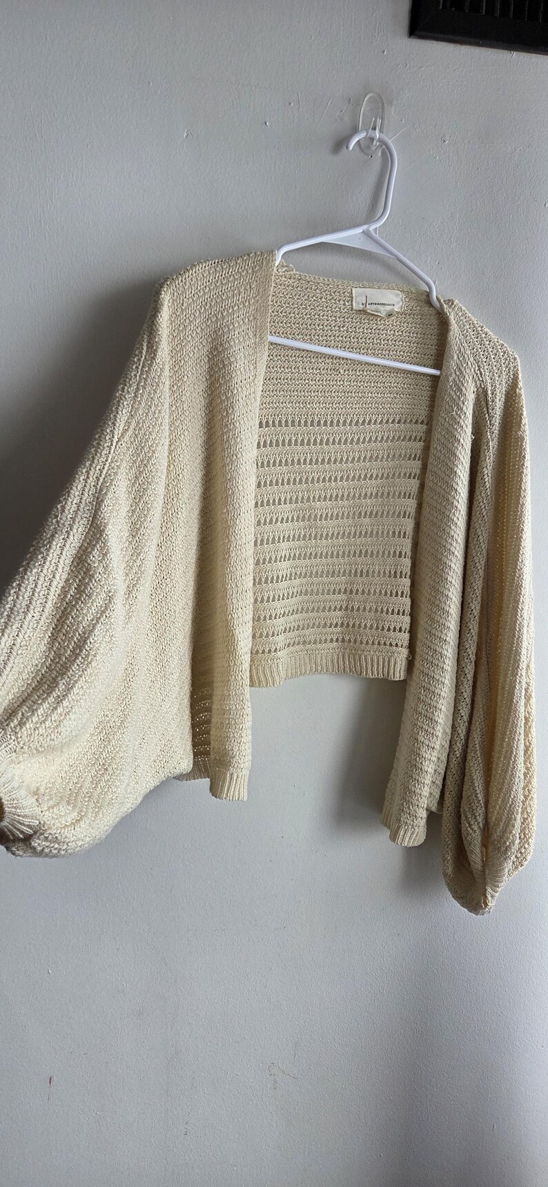 May include: Cream-colored open-front cardigan with long, voluminous sleeves. The knitwear features a textured pattern on the sleeves and a more open, airy weave on the body. The garment is hanging on a white hanger against a plain wall.