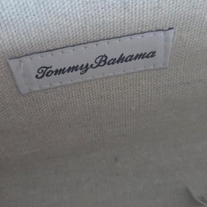 Puede incluir: El interior de un bolso con un cierre de marco met&aacute;lico dorado. Una etiqueta de tela blanca rectangular est&aacute; cosida en el forro de lona texturizada de color blanco roto, mostrando el nombre de la marca Tommy Bahama en letras cursivas negras.