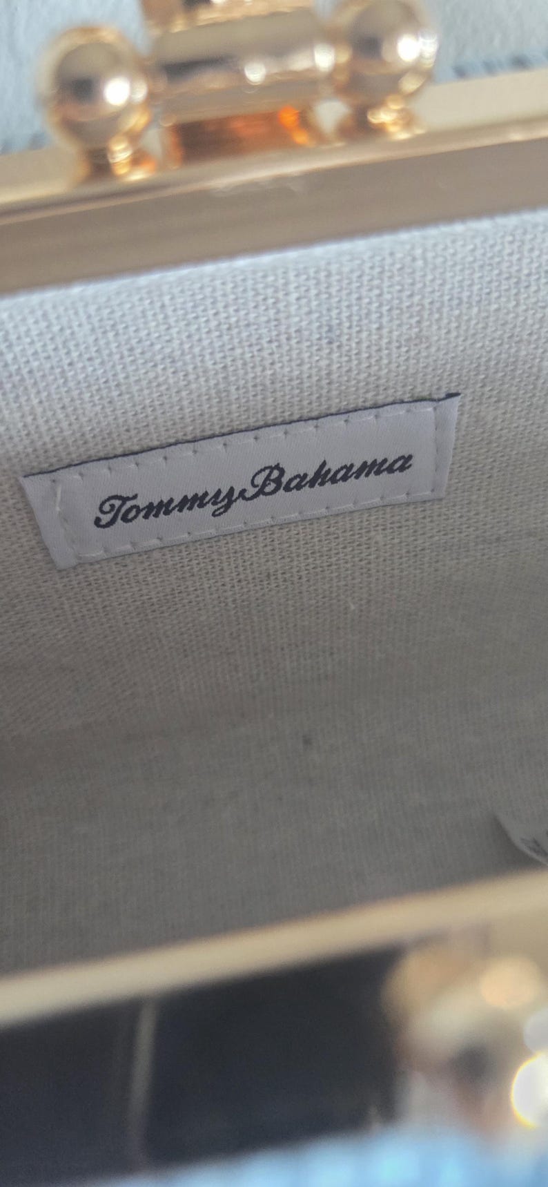 Puede incluir: El interior de un bolso con un cierre de marco met&aacute;lico dorado. Una etiqueta de tela blanca rectangular est&aacute; cosida en el forro de lona texturizada de color blanco roto, mostrando el nombre de la marca Tommy Bahama en letras cursivas negras.