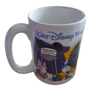 May include: White ceramic mug featuring the words "Walt Disney World" in blue script. The mug has a handle and a colorful graphic of Disney characters, including Donald Duck and Daisy Duck, with the text "Disney-MGM Studios".