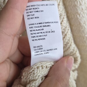 May include: Cream-colored knitwear with a textured pattern. A tag with care instructions is visible. The tag includes washing instructions, such as "wash cold with like colors" and "do not bleach."