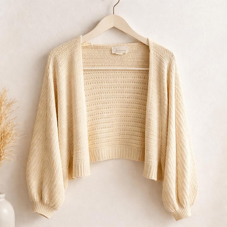 May include: Cream-colored open-front cardigan with long, voluminous sleeves. The knitwear features a textured pattern and is displayed on a wooden hanger. The cardigan is a neutral tone, suitable for layering.