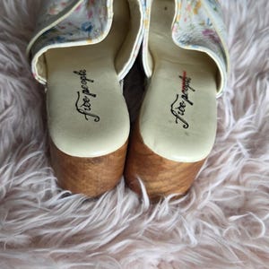 May include: Pair of clogs with floral print uppers and wooden soles. The clogs have a cream-colored base with a colorful floral pattern. The soles are made of light brown wood. The brand name "Free People" is printed on the insole.