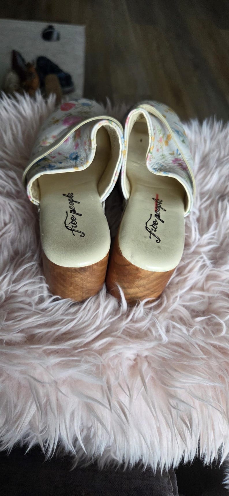 May include: Pair of clogs with floral print uppers and wooden soles. The clogs have a cream-colored base with a colorful floral pattern. The soles are made of light brown wood. The brand name "Free People" is printed on the insole.
