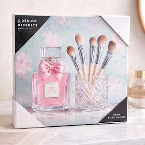Puede incluir: Un juego de regalo con un frasco de perfume rosa con un lazo rosa, un tarro de cristal transparente con brochas de maquillaje y una caja con el texto "DESIGN DISTRICT". El fondo tiene un estampado floral.