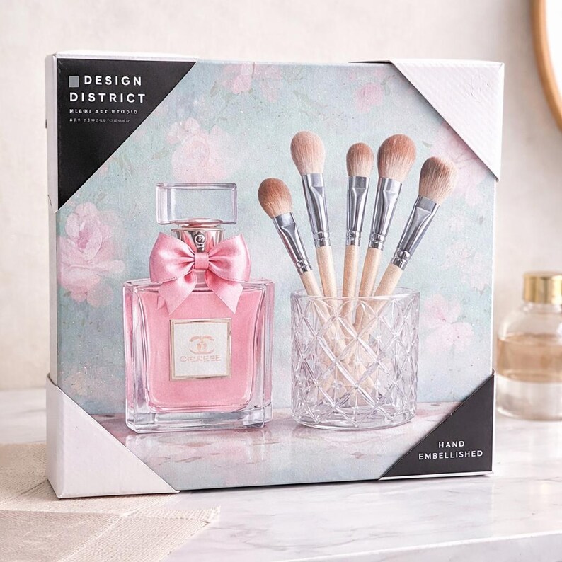 Puede incluir: Un juego de regalo con un frasco de perfume rosa con un lazo rosa, un tarro de cristal transparente con brochas de maquillaje y una caja con el texto "DESIGN DISTRICT". El fondo tiene un estampado floral.