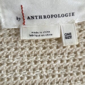 May include: Close-up of a cream-colored knit fabric with a tag that reads "by ANTHROPOLOGIE." The tag also states "made in china" and "ONE SIZE."
