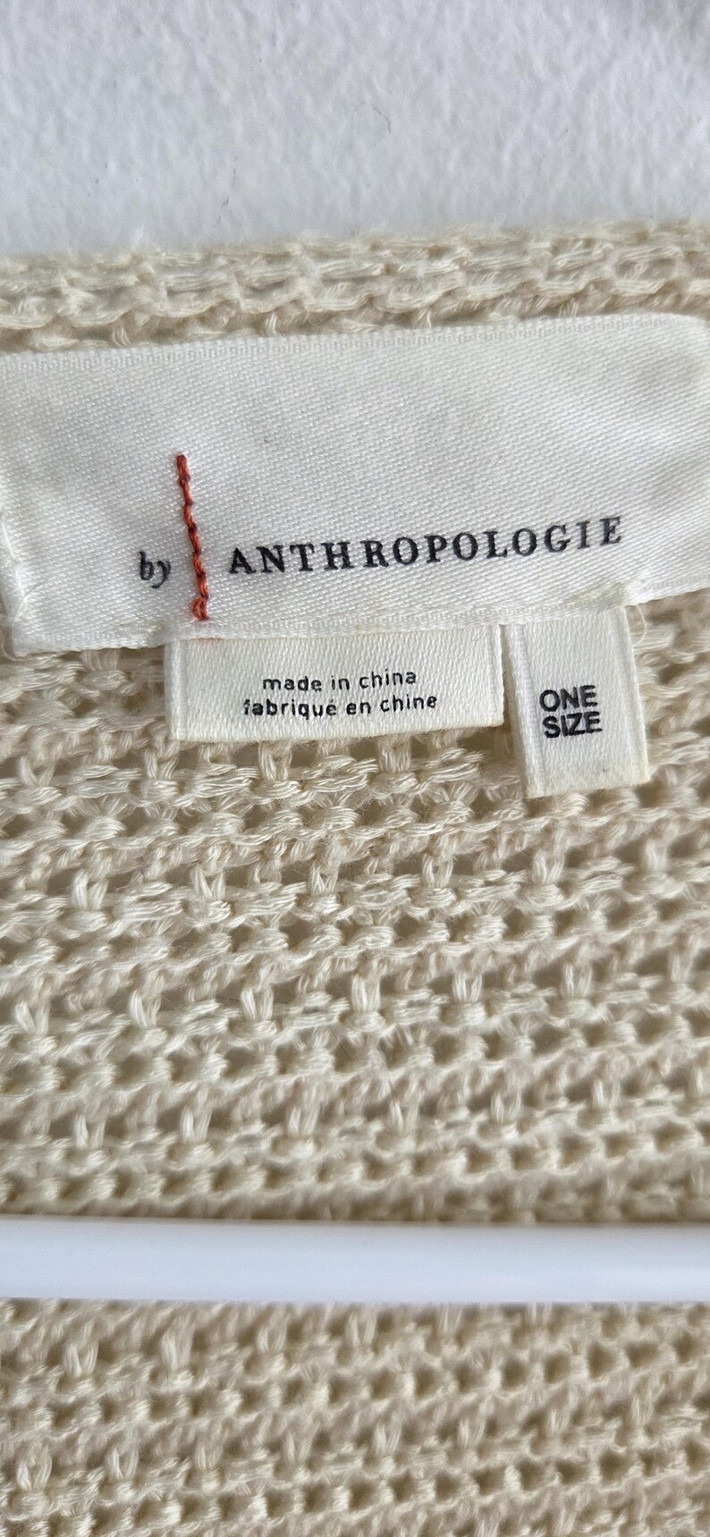 May include: Close-up of a cream-colored knit fabric with a tag that reads "by ANTHROPOLOGIE." The tag also states "made in china" and "ONE SIZE."