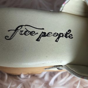 May include: Close-up of the interior of a shoe with the words "Free People" in black script. The shoe's insole is a light cream color, and the sole is made of wood. The background is a soft, fluffy, light pink material.