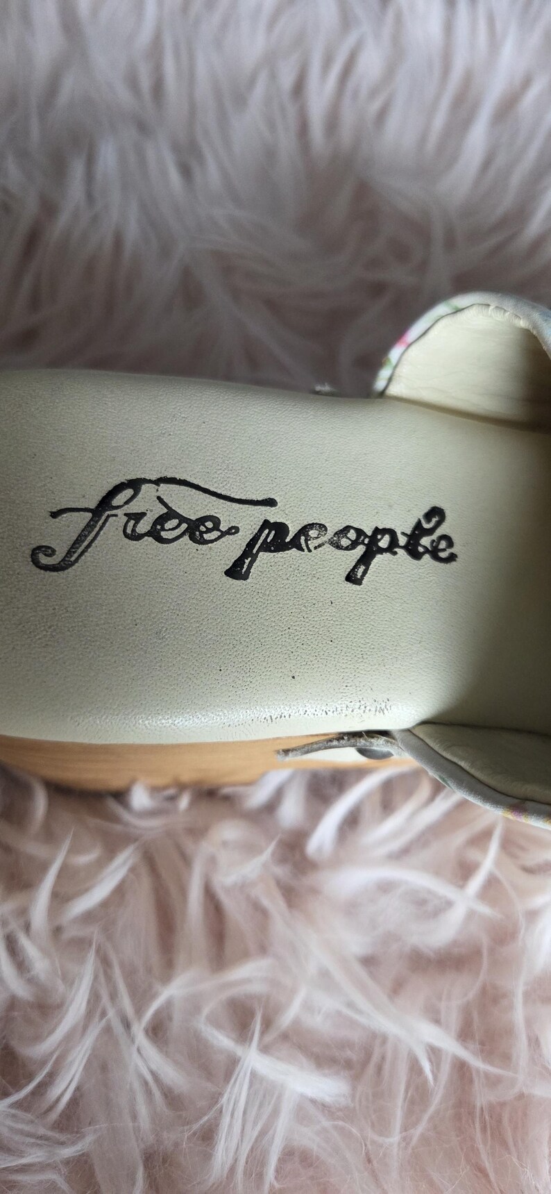 May include: Close-up of the interior of a shoe with the words "Free People" in black script. The shoe's insole is a light cream color, and the sole is made of wood. The background is a soft, fluffy, light pink material.