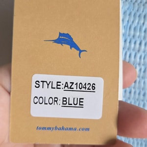 Puede incluir: Una etiqueta de producto rectangular de color tostado con un logotipo de marl&iacute;n azul, el texto STYLE:AZ10426, COLOR:BLUE y tommybahama.com. La etiqueta est&aacute; unida a un art&iacute;culo azul claro texturizado con una cadena dorada, visible a la derecha.