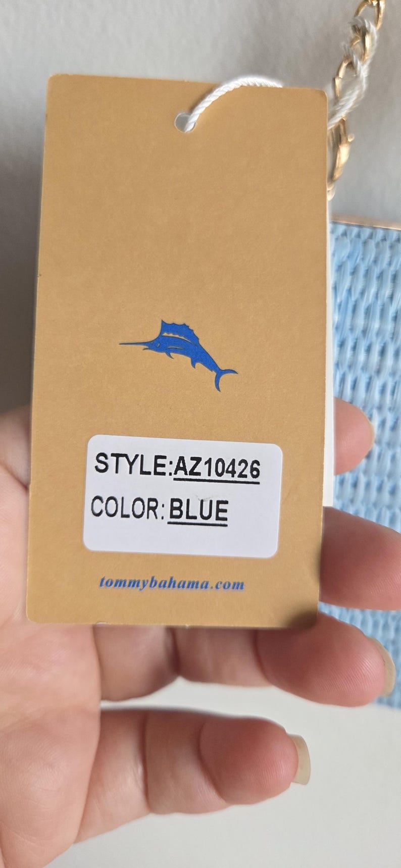 Puede incluir: Una etiqueta de producto rectangular de color tostado con un logotipo de marl&iacute;n azul, el texto STYLE:AZ10426, COLOR:BLUE y tommybahama.com. La etiqueta est&aacute; unida a un art&iacute;culo azul claro texturizado con una cadena dorada, visible a la derecha.