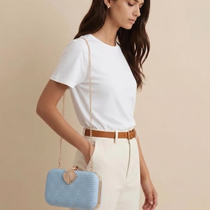 Puede incluir: Un bolso de mano rectangular de color azul claro con una superficie tejida texturizada, que presenta un marco de metal dorado y un cierre decorativo en forma de concha. El bolso incluye una fina correa de cadena dorada y bordes met&aacute;licos.