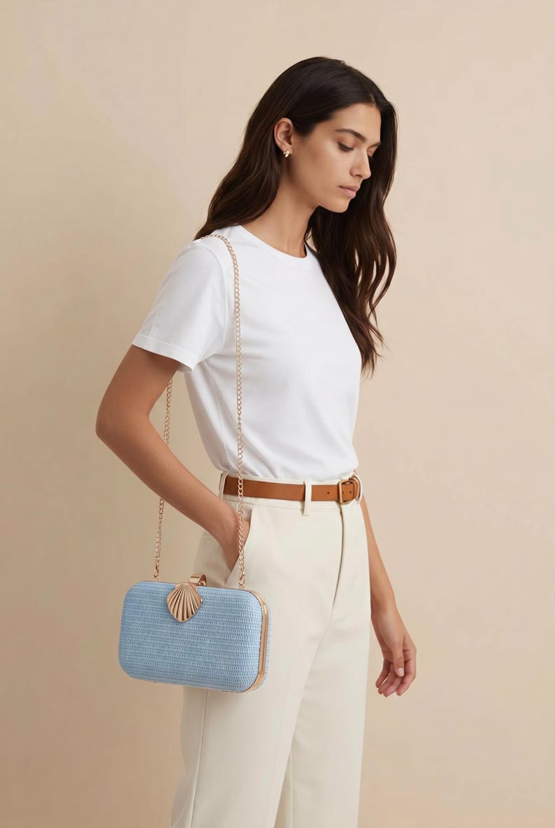 Puede incluir: Un bolso de mano rectangular de color azul claro con una superficie tejida texturizada, que presenta un marco de metal dorado y un cierre decorativo en forma de concha. El bolso incluye una fina correa de cadena dorada y bordes met&aacute;licos.
