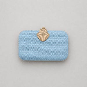 Puede incluir: Un bolso de mano rectangular de color azul claro con textura tejida. El bolso presenta un cierre de metal dorado con forma de concha marina centrado en el borde superior. El marco del bolso tambi&eacute;n est&aacute; acabado en un material met&aacute;lico dorado a juego.