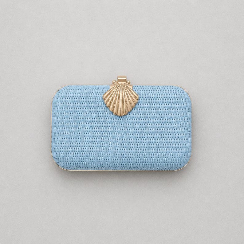 Puede incluir: Un bolso de mano rectangular de color azul claro con textura tejida. El bolso presenta un cierre de metal dorado con forma de concha marina centrado en el borde superior. El marco del bolso tambi&eacute;n est&aacute; acabado en un material met&aacute;lico dorado a juego.