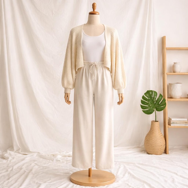 May include: A cream-colored outfit on a mannequin. The outfit includes a cropped cardigan with balloon sleeves, a white tank top, and wide-leg pants. The outfit is displayed on a wooden mannequin stand.