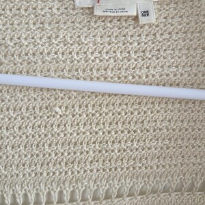 May include: Cream-colored open-knit cardigan with a tag that reads "by ANTHROPOLOGIE". The sweater is made of a textured, open weave fabric and is hanging on a white hanger. The tag also says "ONE SIZE".
