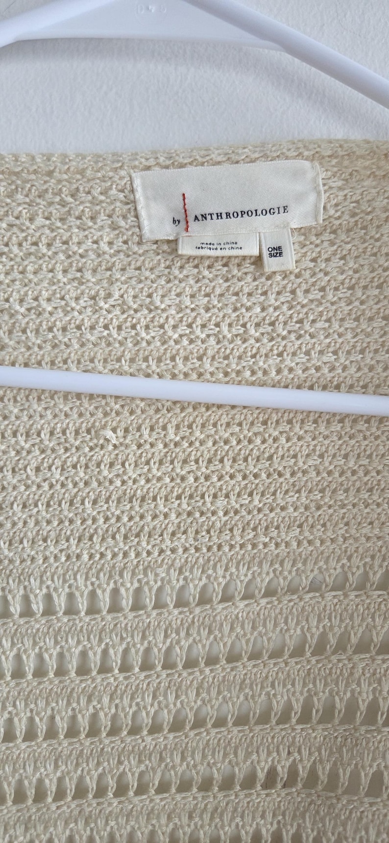 May include: Cream-colored open-knit cardigan with a tag that reads "by ANTHROPOLOGIE". The sweater is made of a textured, open weave fabric and is hanging on a white hanger. The tag also says "ONE SIZE".