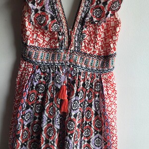 May include: A patterned summer dress with a deep V-neck and ruffled sleeves. The dress features a floral design in red, black, and purple, with a tassel detail. The dress is approximately 100 cm in length.