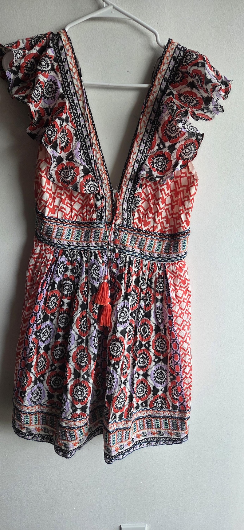 May include: A patterned summer dress with a deep V-neck and ruffled sleeves. The dress features a floral design in red, black, and purple, with a tassel detail. The dress is approximately 100 cm in length.