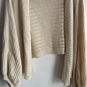 May include: Cream-colored open-front cardigan with long, voluminous sleeves. The knitwear features a textured pattern and is displayed on a white hanger against a white wall. A tag is visible on the back of the neck.